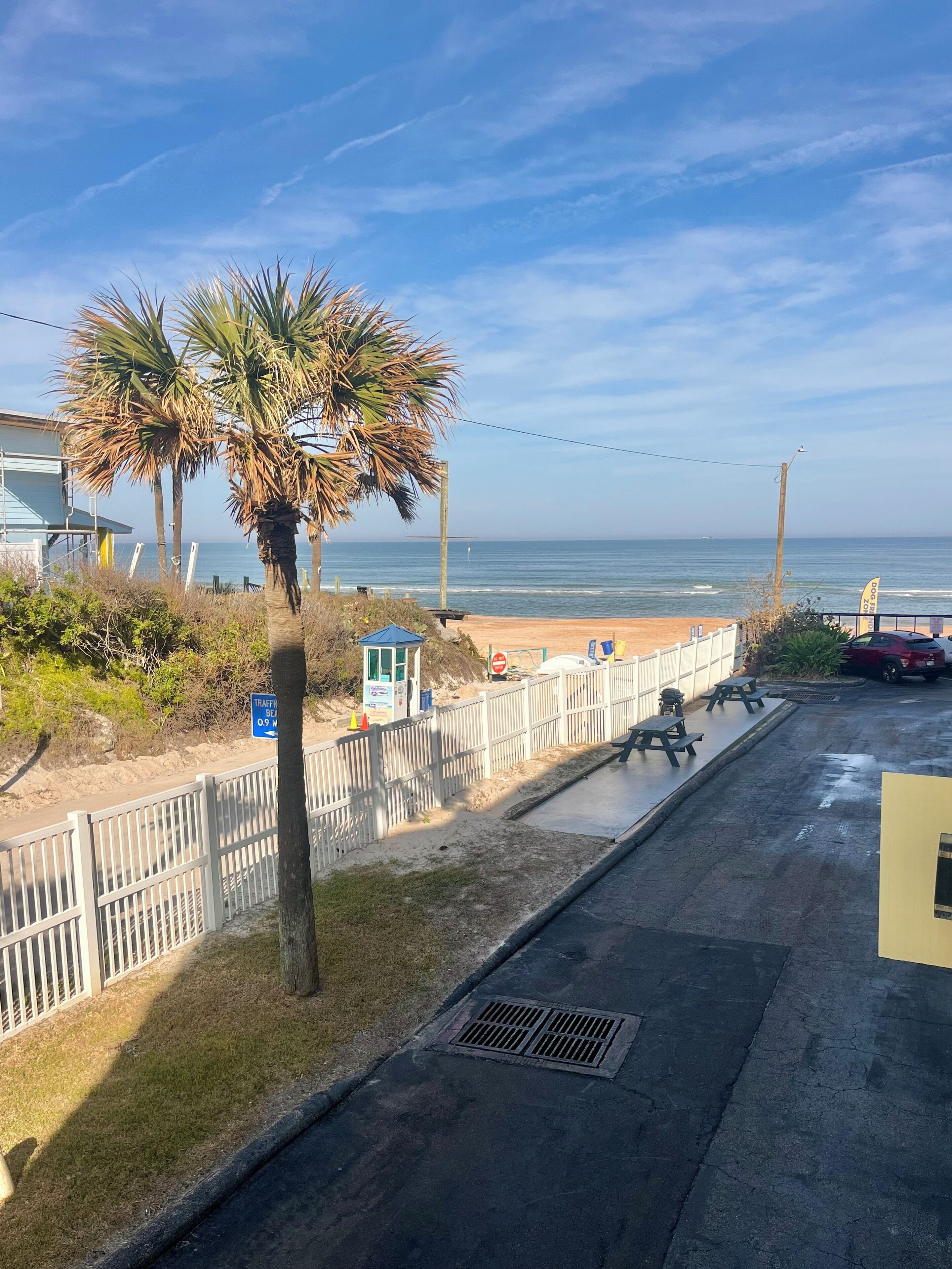 Eine Küstenstraße mit Blick auf einen Strand, Palmen, Picknicktischen und einem Rettungsturm.