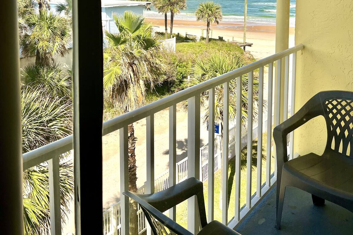A balcony with a view of the beach, palm trees, and a clear sky.