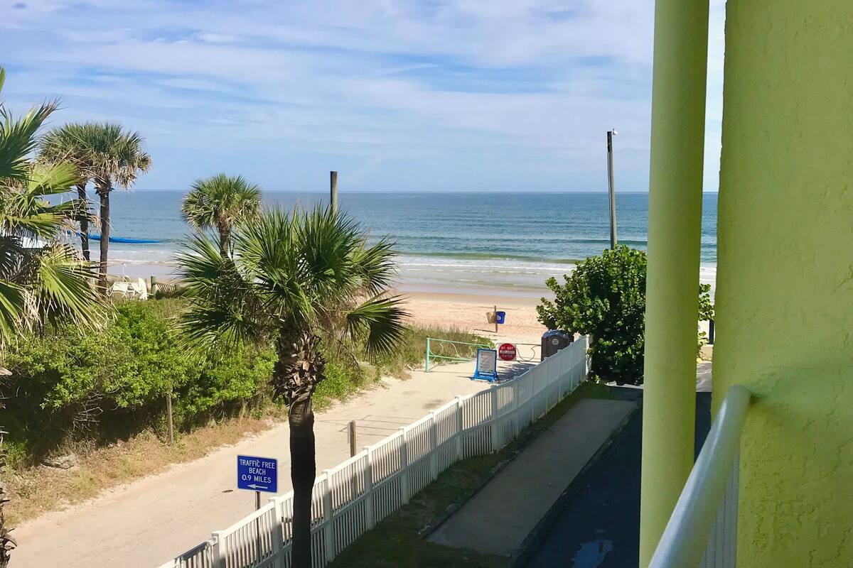 A beach view with a white fence, palm trees, and a sign indicating a traffic-free beach 0.9 miles away.