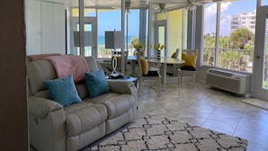 A living room with a beige sofa, a coffee table, and a dining area with chairs and a table. There is a large window with a view of the ocean and buildings. - Symphony Beach Club (Ormond Beach)