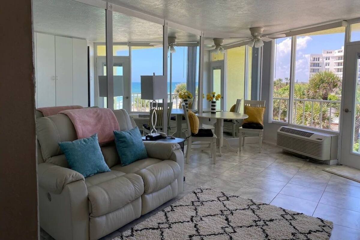 A living room with a beige sofa, a coffee table, and a dining area with chairs and a table. There is a large window with a view of the ocean and buildings.