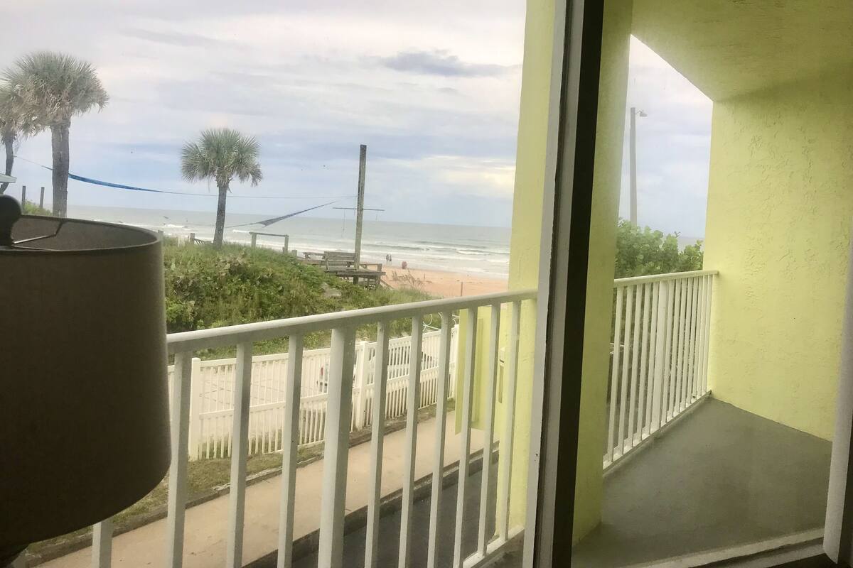 A balcony with a view of the beach, white railing, and a lamp.