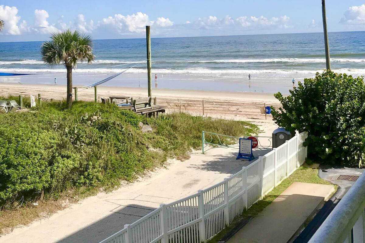 A beach view with a white fence, a hammock, and a palm tree.