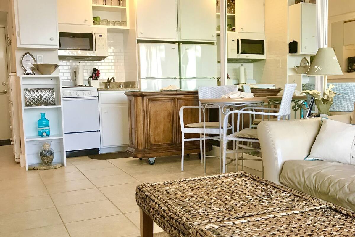 A modern kitchen with white cabinets, a microwave, and a stove. There is a dining area with a wicker table and chairs, and a sofa in the living room.