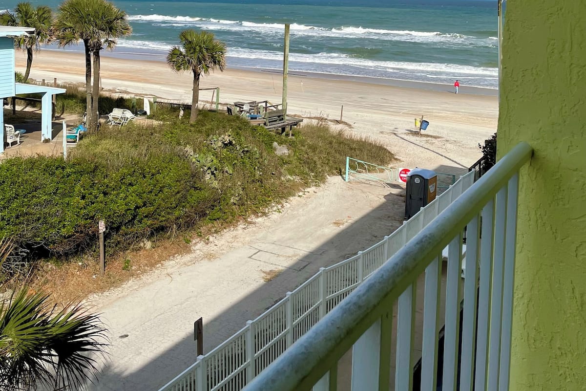 A beach view from a balcony with a clear sky, a sandy beach, and a few palm trees.