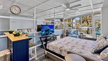 Een moderne slaapkamer met een bed, een bureau, een televisie en een groot raam met uitzicht op palmbomen.