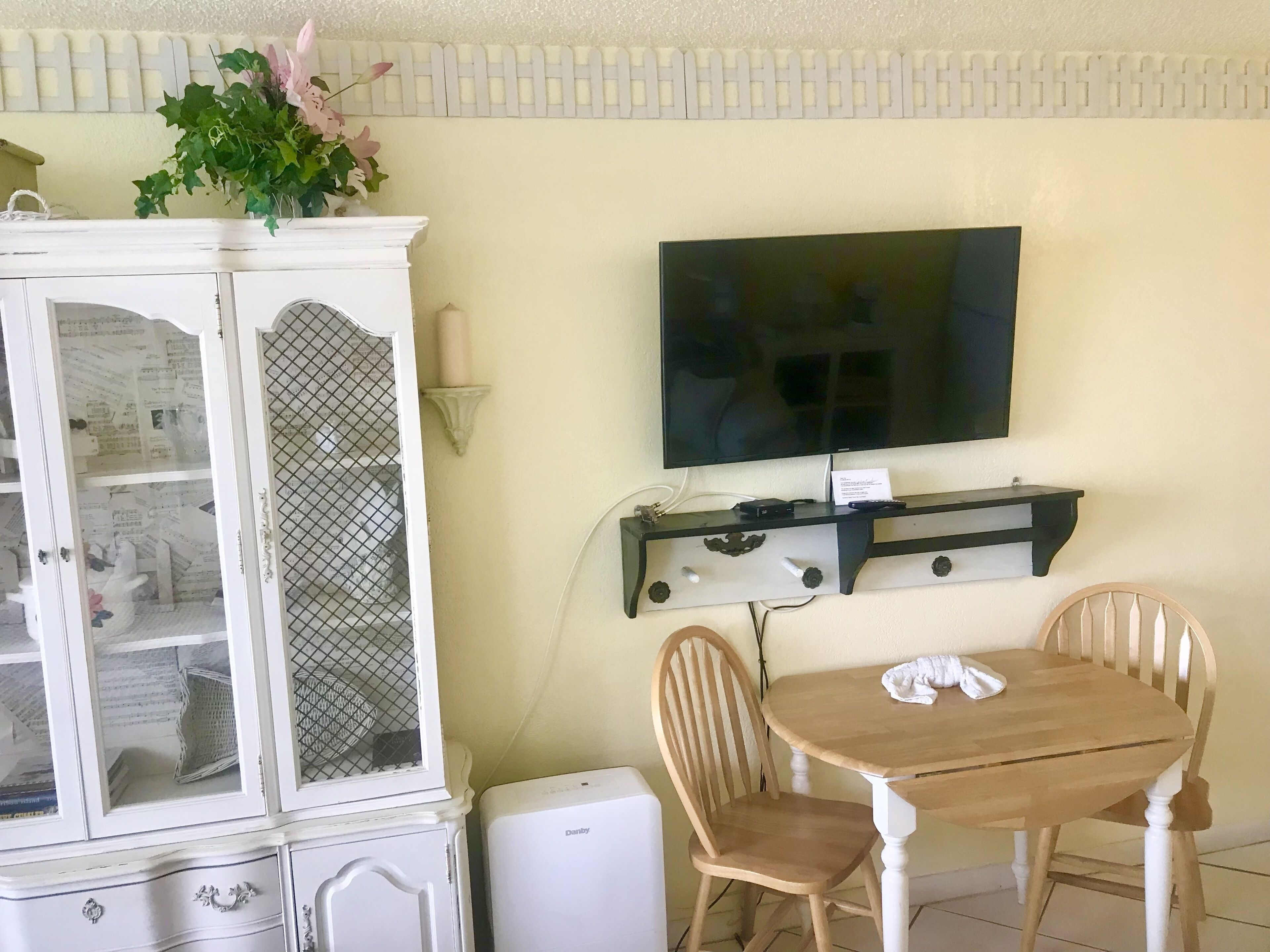 Un comedor con una mesa y sillas de madera, un armario blanco con puertas de cristal y un televisor de pantalla plana montado en la pared.