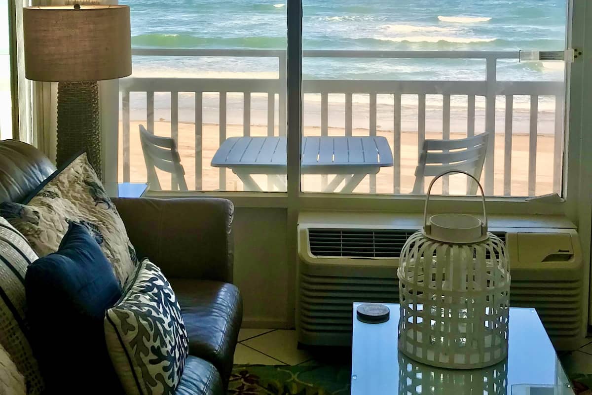 A room with a view of the ocean, a sofa with patterned cushions, a table with a lantern, and a chair.