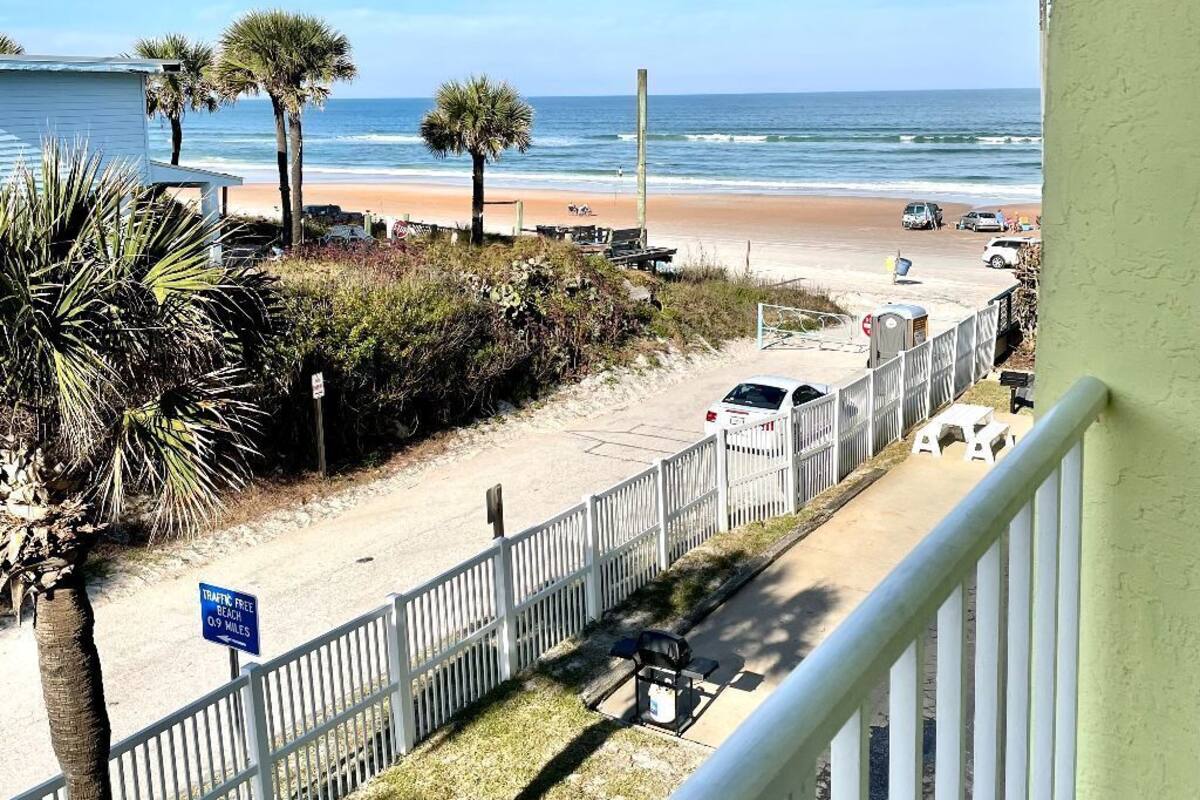 A beachfront view with a white fence, palm trees, and a beach in the background.