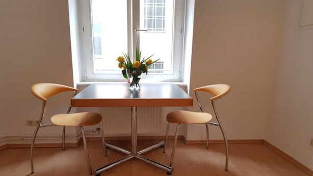 A dining area with a table and two chairs, a window with a view, and a vase with flowers on the table.