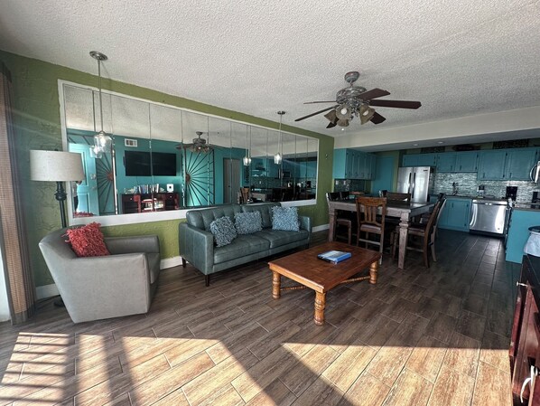 A modern living room with a sofa, coffee table, and a dining area with chairs. A kitchen with blue cabinets and stainless steel appliances is visible.