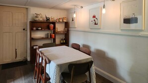 A dining area with a table and chairs, a wooden shelf with kitchenware, and framed artwork on the wall.