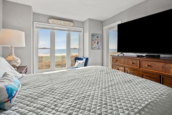 A bedroom with a large bed, a TV, a dresser, and a view of the beach through a large window.