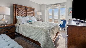 Ein Schlafzimmer mit einem großen Bett, einem Schreibtisch, einem Stuhl und Blick auf den Strand durch das Fenster.