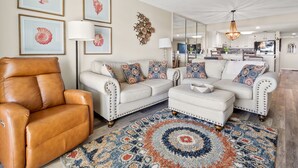 Ein Wohnzimmer mit einem braunen Ledersessel, einem beige-farbenen Sofa mit gemusterten Kissen, einem passenden Hocker und einem großen Spiegel, der den Küchenbereich reflektiert.
