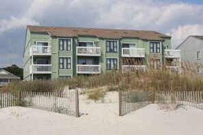A multi-story building with balconies, situated on a sandy beach with a wooden fence in the foreground.