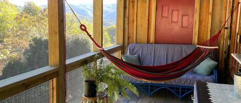 A wooden deck with a hammock, a bed with a checkered bedspread, and a view of the outdoors.