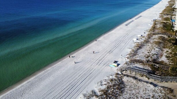Ovenudblik over en strand med et langt sandstykke, et klart blåt hav og et par personer der går.