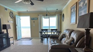 En stue med en lædersofa, et spisebord med stole, en loftsvift og udsigt til havet gennem en skydedør.