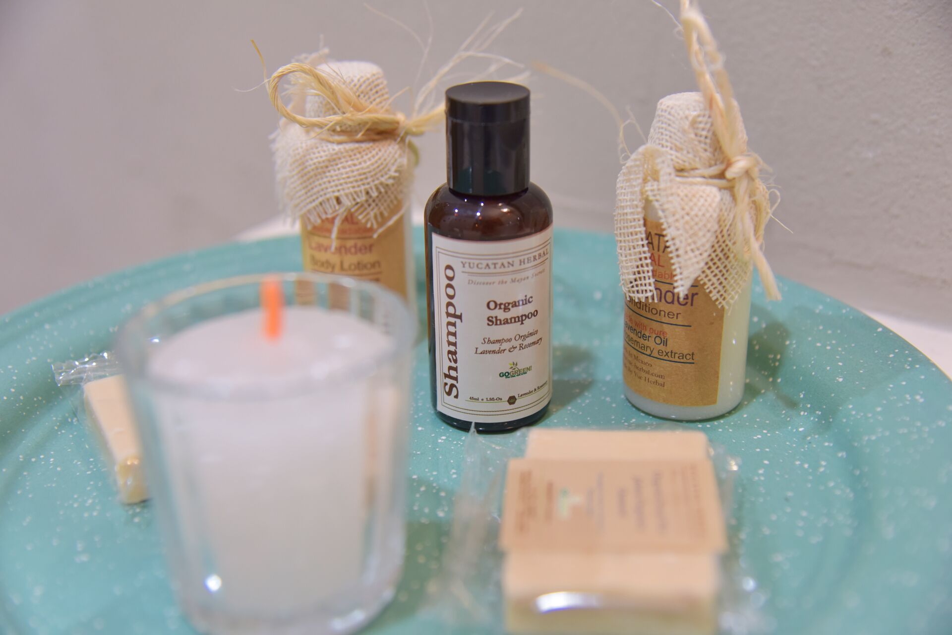 Un vassoio color petrolio con una candela, una bottiglia di shampoo biologico e due piccoli vasetti di lozione corpo.