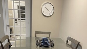 Un comedor con una mesa de vidrio, cuatro sillas, un reloj en la pared y una puerta que da a otra habitación.