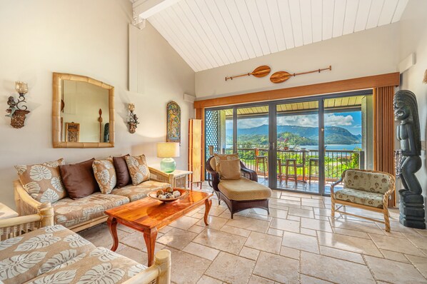 A spacious living area with a sofa, chairs, a coffee table, and a view of a mountainous landscape through large sliding doors. - (4323/24) 2 Br Hanalei Bay Resort with Amazing Ocean View (Princeville)