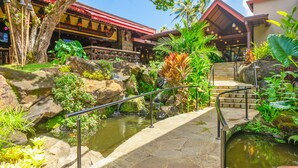 A tropical resort with a stone bridge over a pond, surrounded by lush greenery and a restaurant with a red roof. - (4323) 1 Br Hanalei Bay Resort, Ocean view and Plenty of Amenities (Princeville)