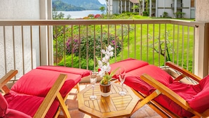 A balcony with wooden chairs and red cushions, a small table with a potted plant and two glasses, and a view of a lake and buildings in the background. - (4324) Studio at Hanalei Bay Resort, Ocean view, Pool (Princeville)