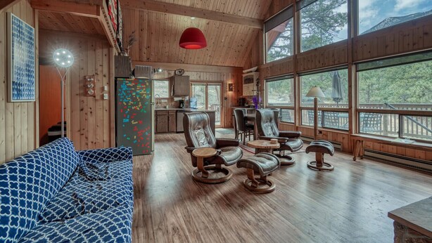 Un interior acogedor de cabaña con detalles de madera, una zona de comedor y ventanas amplias que ofrecen vistas al exterior.