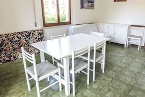 A dining area with a white table and chairs, a bench with a floral pattern, and a window with a view of greenery.