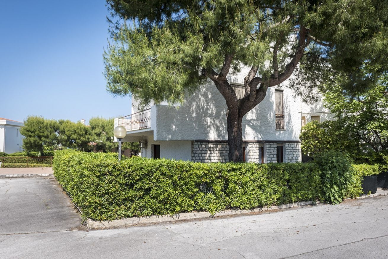 Un edificio di due piani con un albero di fronte, un siepe e un cielo sereno.