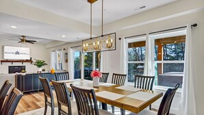 Un espace repas comprenant une grande table, des chaises, un lustre et une vue sur l’extérieur grâce à de grandes fenêtres.
