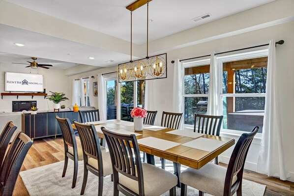 A dining area with a large table, chairs, a chandelier, and a view of the outdoors through large windows. - Enjoy Arcade Games! Ski Ball, Shuffleboard, Air Hockey, Dart Board, Hot Tub & Cmty Pools (Hollister)