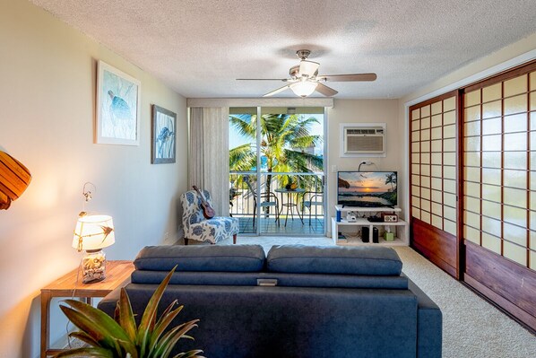 A living room with a blue sofa, a ceiling fan, a lamp, and a view of palm trees through large glass doors.