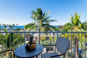 A balcony with a table and chairs, a bowl of fruit, and a view of the ocean and palm trees.