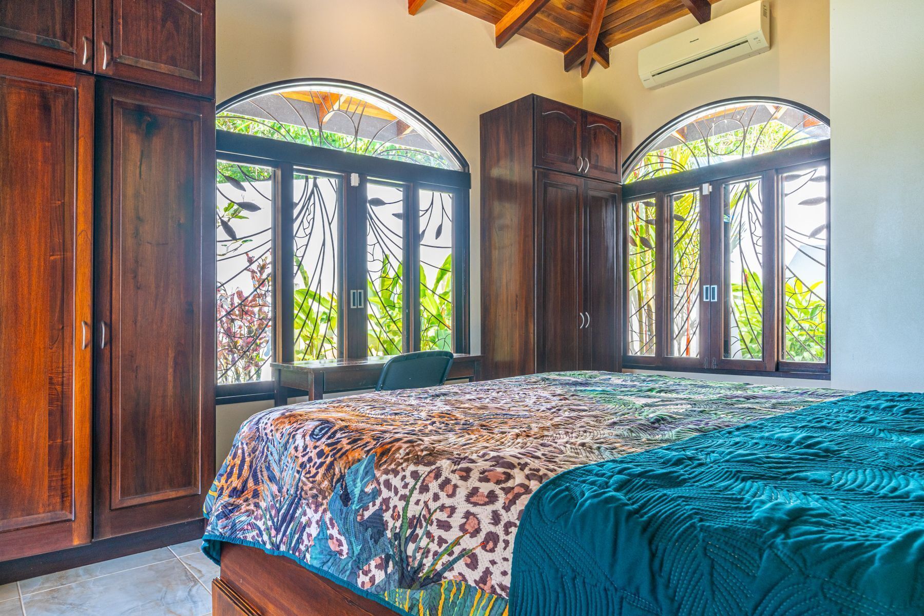 Un dormitorio con una cama grande, un armario de madera y dos ventanas arqueadas con vistas a zonas verdes.
