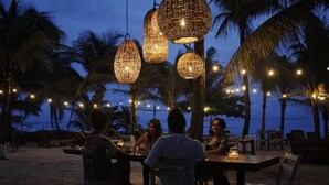 An outdoor dining area with wicker hanging lights and palm trees, overlooking a beach. - Beachfront 1BR Apartment w/ Terrace & Beach Club (Mahahual)