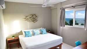 A bedroom with a bed, a ceiling fan, a window with a view of the ocean, a wooden nightstand, and a small potted plant. - Beachfront 1BR Apartment w/ Terrace & Beach Club (Mahahual)