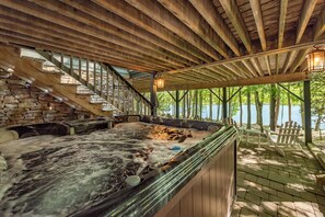 A hot tub area with stone walls and wooden beams, surrounded by a covered deck with Adirondack chairs. - Lakefront Luxury w/ Fireplaces, Hot Tub & Dock (McHenry)