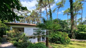 A house with a paved driveway, surrounded by lush greenery and tall trees. - Attractive family home for 10 people in popular Hossegor lake district (SOORTS HOSSEGOR)