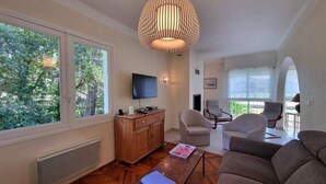 A spacious living room with a large window, a wooden cabinet, a television, and a modern hanging light fixture. - Attractive family home for 10 people in popular Hossegor lake district (SOORTS HOSSEGOR)