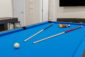 A pool table with two cues and a white cue ball, a set of colorful pool balls, and a television mounted on the wall. - Unique Lakeview Home with Indoor Pool in Basement! (Branson)