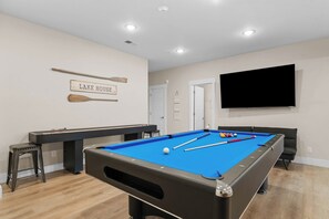 Une table de billard recouverte d’un drap bleu, un queue de billard et des billes. Un téléviseur est fixé au mur, et une pancarte indique « LAKE HOUSE ».
