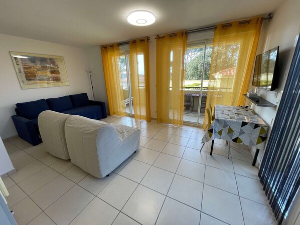 A living room with a sofa, a dining table with chairs, a TV, and a sliding glass door with curtains.