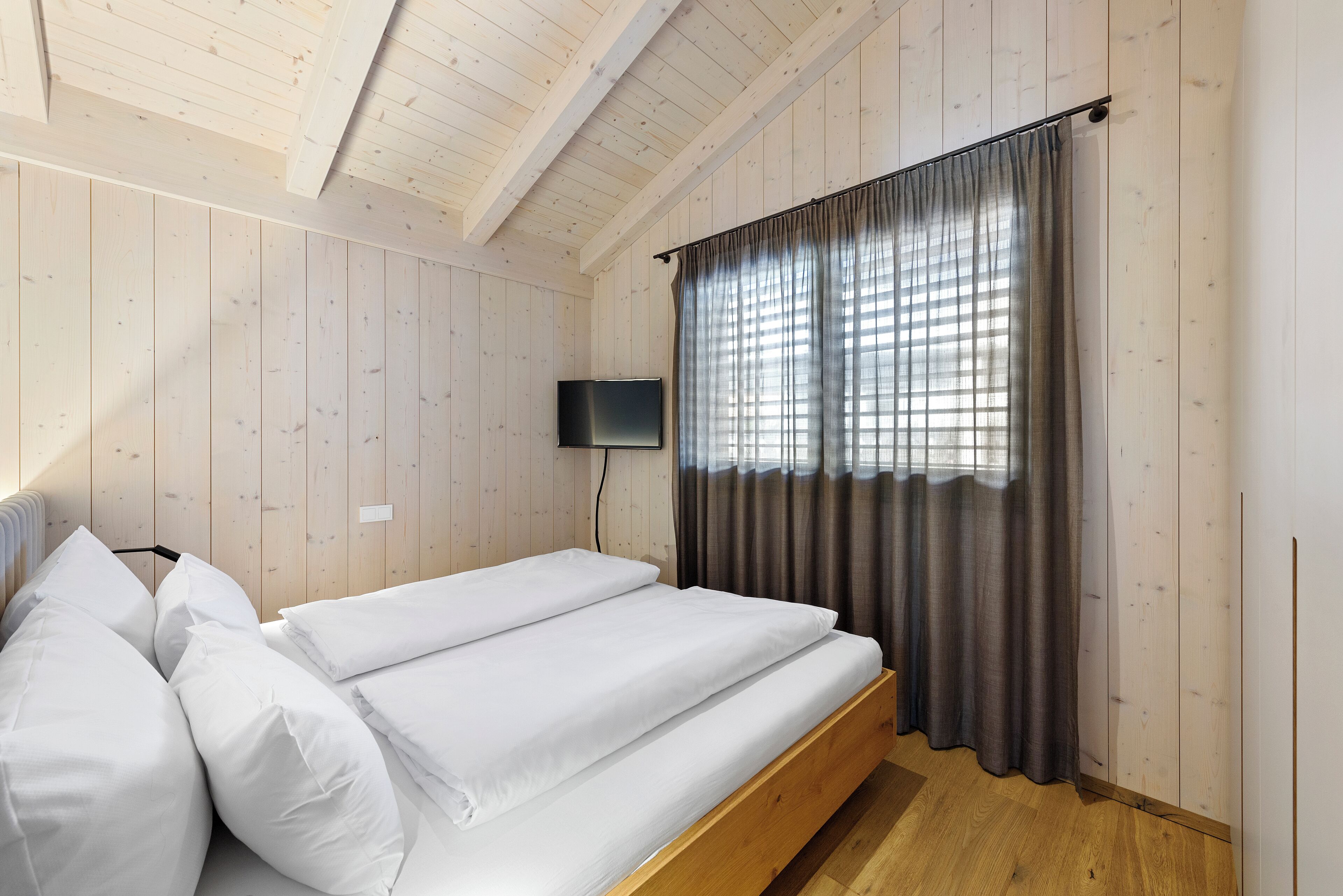 A room with a wooden ceiling, a bed with white bedding, a TV mounted on the wall, and a window with curtains.