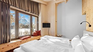 Un dormitorio con un ventanal grande, vigas de madera, una cama, un televisor y una mesita de noche.
