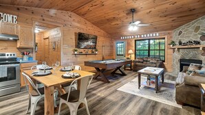A cozy cabin interior with a wooden dining table, a pool table, a fireplace, and a stone wall.