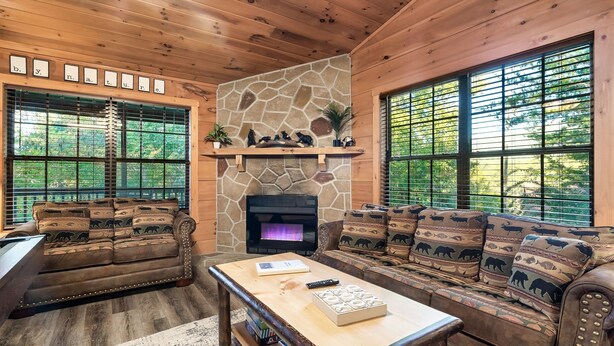 A cozy living room with a stone fireplace, two sofas with patterned cushions, a coffee table, and large windows with blinds.