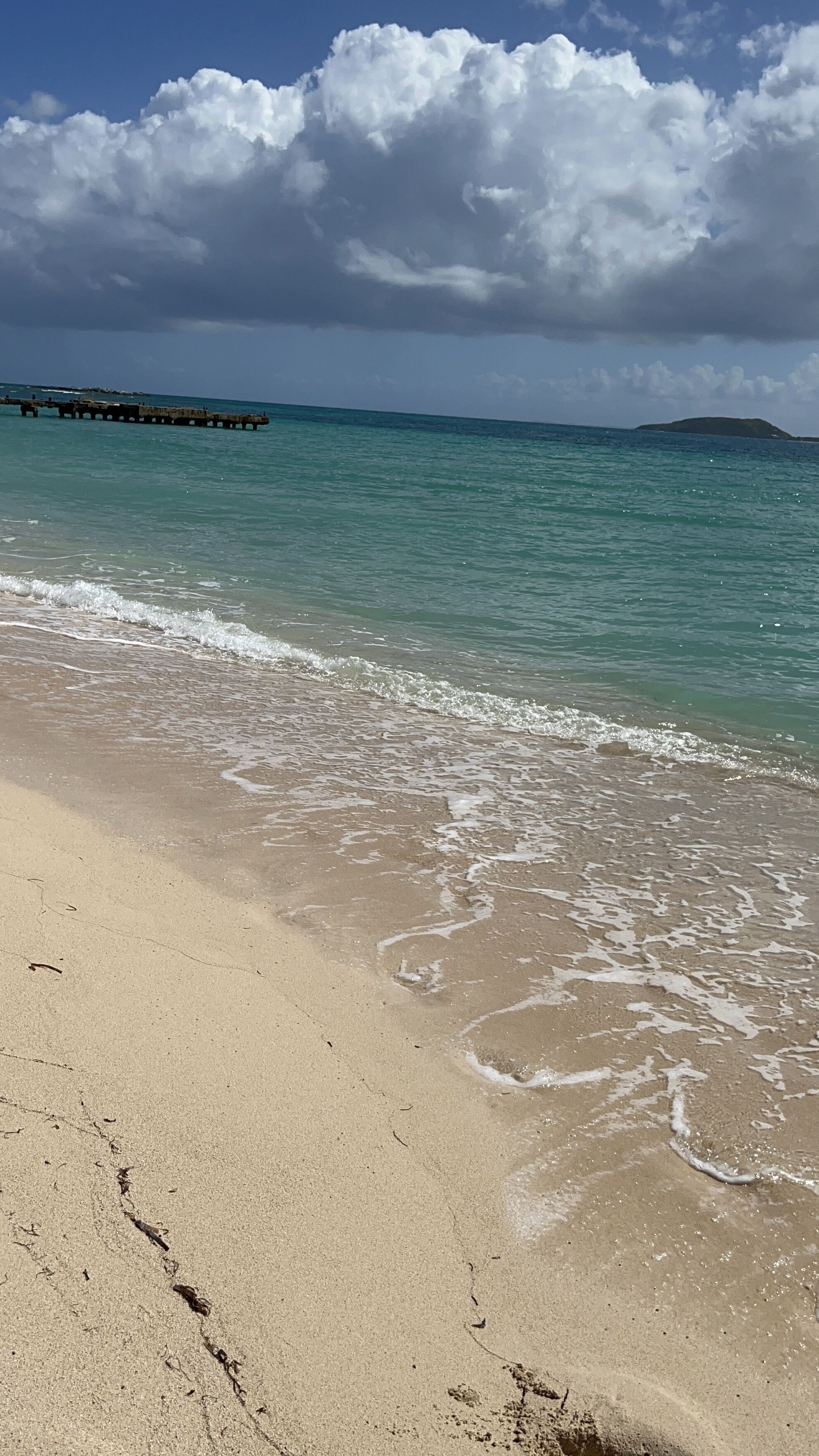 A beach with clear turquoise water, a sandy shore, and a distant pier.