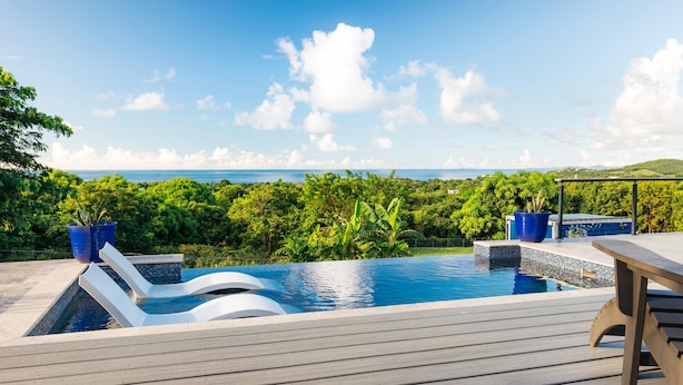 A swimming pool with a wooden deck, surrounded by lush greenery and a view of the ocean. - Tiny house on an acre with ocean view and private infinity pool.  (Vieques)